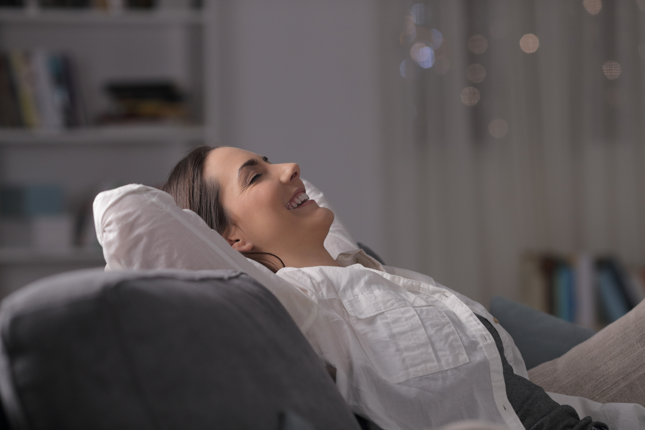 A woman sat on a sofa with her hands behind her head smiling.