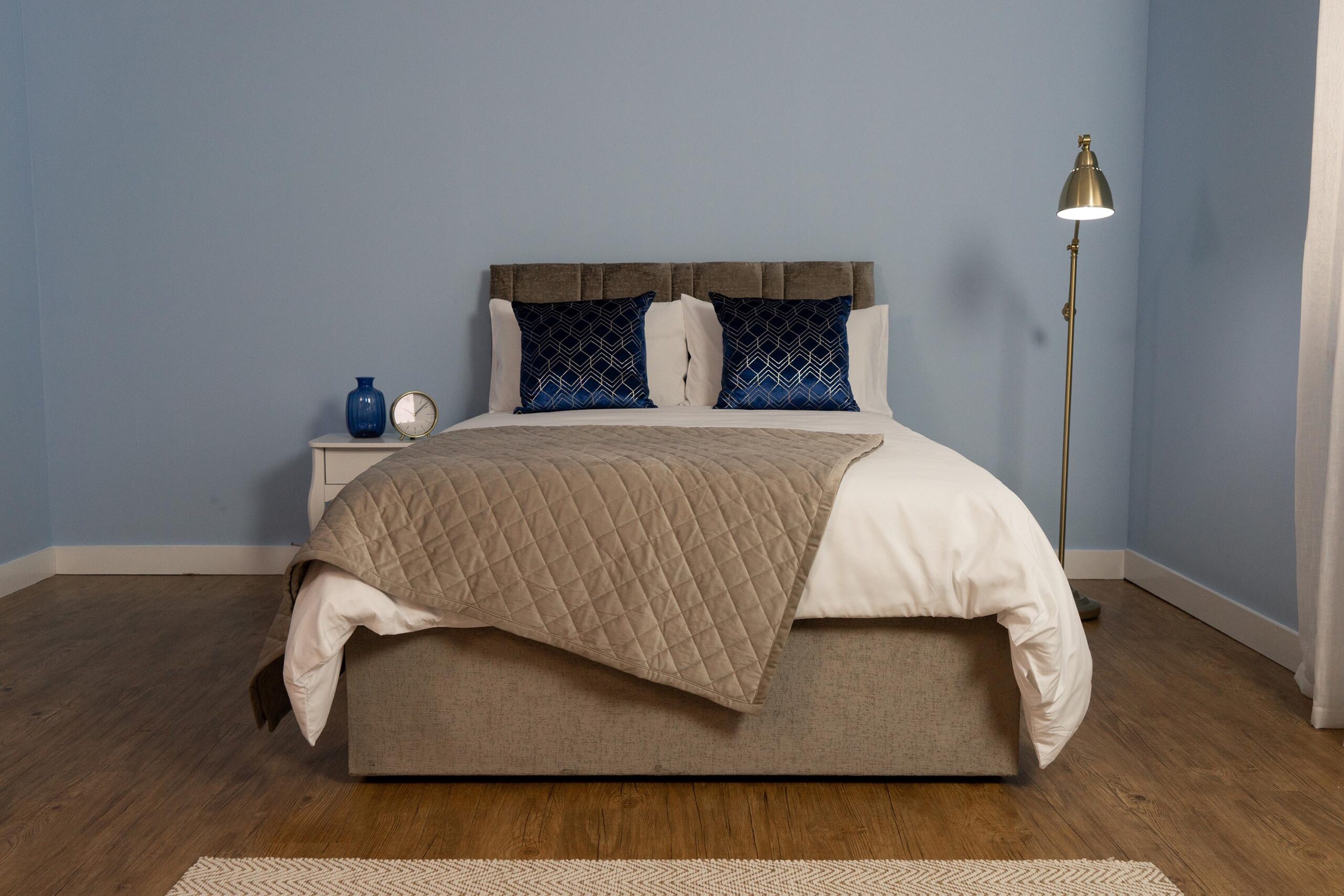 The Majestic Adjustable bed with beige fabric, white sheets, blue cushions and a beige blanket on top of it.